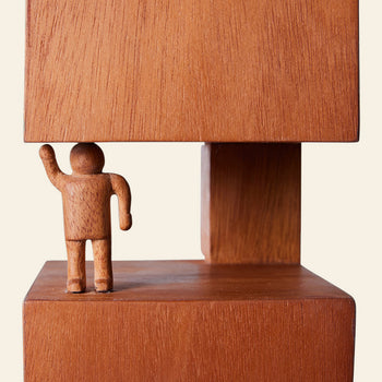 The HKLIVING HK Objects: Empowered is a small wooden figure with one arm raised, standing between two large stacked blocks—an artful sculpture by HKLIVING, ideal for enhancing modern interior spaces.