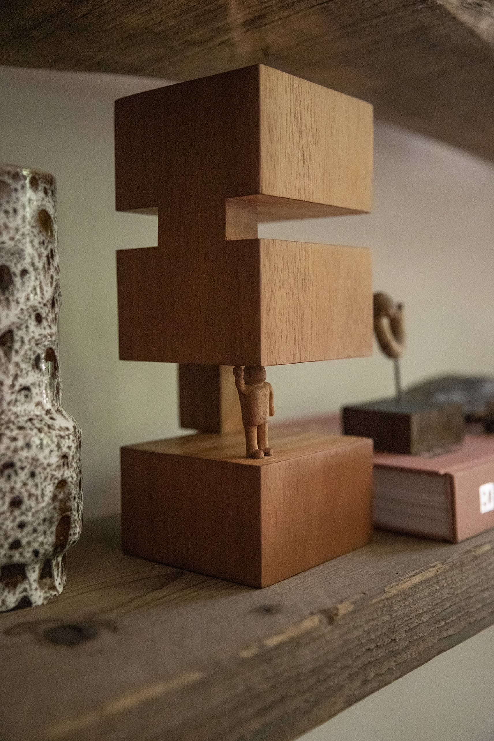 The HKLIVING HK Objects: Empowered, a Bauhaus-inspired small wooden sculpture by HKLIVING, features a figure lifting two rectangular blocks—ideal for modern spaces when displayed with a speckled ceramic vase and a closed book on a rustic shelf.