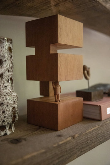 The HKLIVING HK Objects: Empowered, a Bauhaus-inspired small wooden sculpture by HKLIVING, features a figure lifting two rectangular blocks—ideal for modern spaces when displayed with a speckled ceramic vase and a closed book on a rustic shelf.