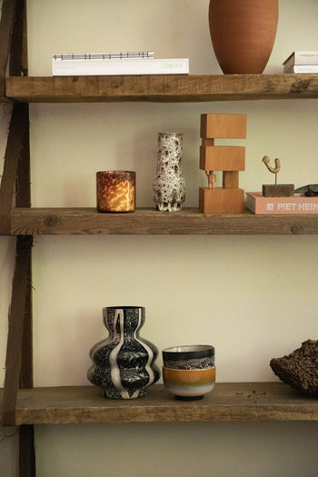 A wooden shelf displays HKLIVING’s Bauhaus-inspired decorative objects from the HK Objects: Empowered collection—including a candle, sculptural vases, stacked wooden blocks, books, and a textured bowl—against a light-colored wall.
