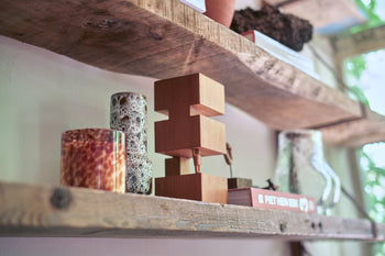 HKLIVING's HK Objects: Empowered display decorative objects on rustic wooden shelves—a speckled glass candle, sculptural art, patterned vase, and book by a sunlit window—perfect accents for modern interiors.