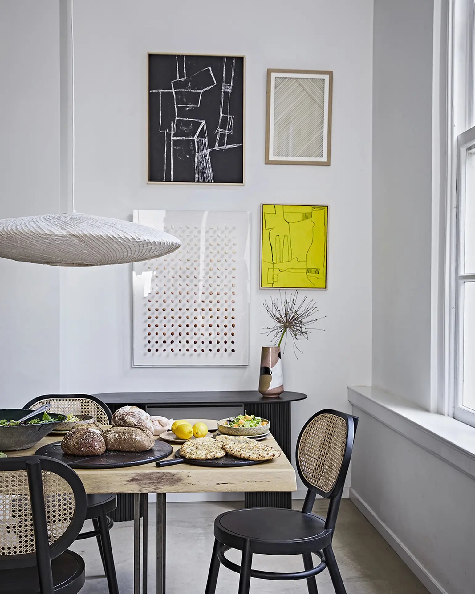A modern dining area features a wooden table set with bread, lemons, and greens. The HKLIVING Hand Painted Ceramic Flower Vase by HKLIVING serves as the centerpiece, while black cane-back chairs and abstract art complete the airy, light-filled space.