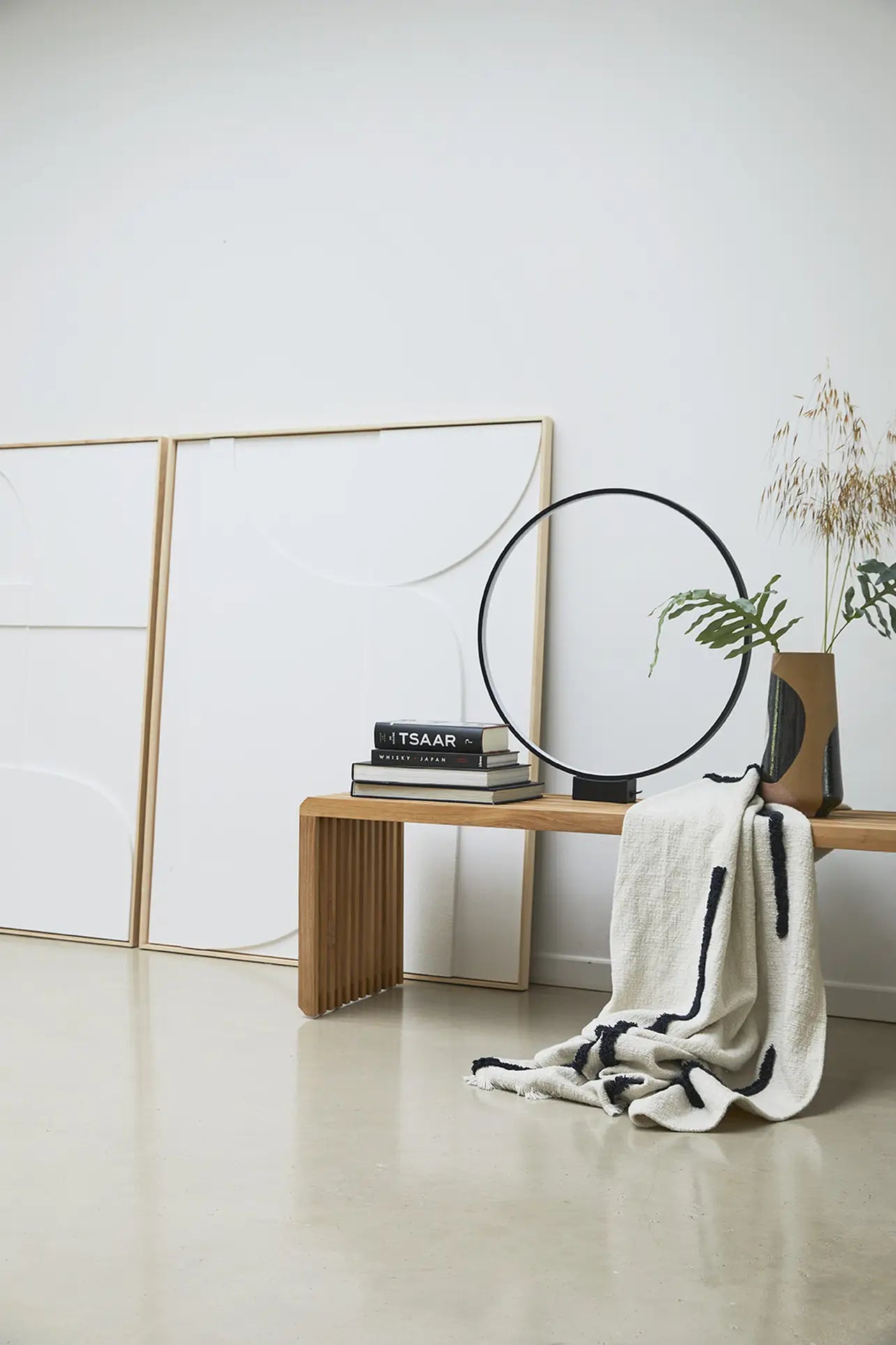 A minimalist interior scene with a wooden bench, black and white books, a round black sculpture, the HKLIVING Hand Painted Ceramic Flower Vase with dried plants, a folded striped blanket, and two blank canvases against a white wall.