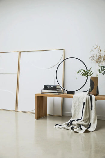 A minimalist interior scene with a wooden bench, black and white books, a round black sculpture, the HKLIVING Hand Painted Ceramic Flower Vase with dried plants, a folded striped blanket, and two blank canvases against a white wall.