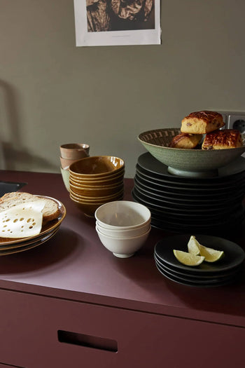 A stack of dark ceramics—plates, bowls, and cups—rests on a maroon surface. Scones fill the HKLIVING Kyoto Ceramics: Japanese Ceramic Salad Bowl, with sliced lime and bread with cheese on a plate. The neutral background shows part of a wall socket.