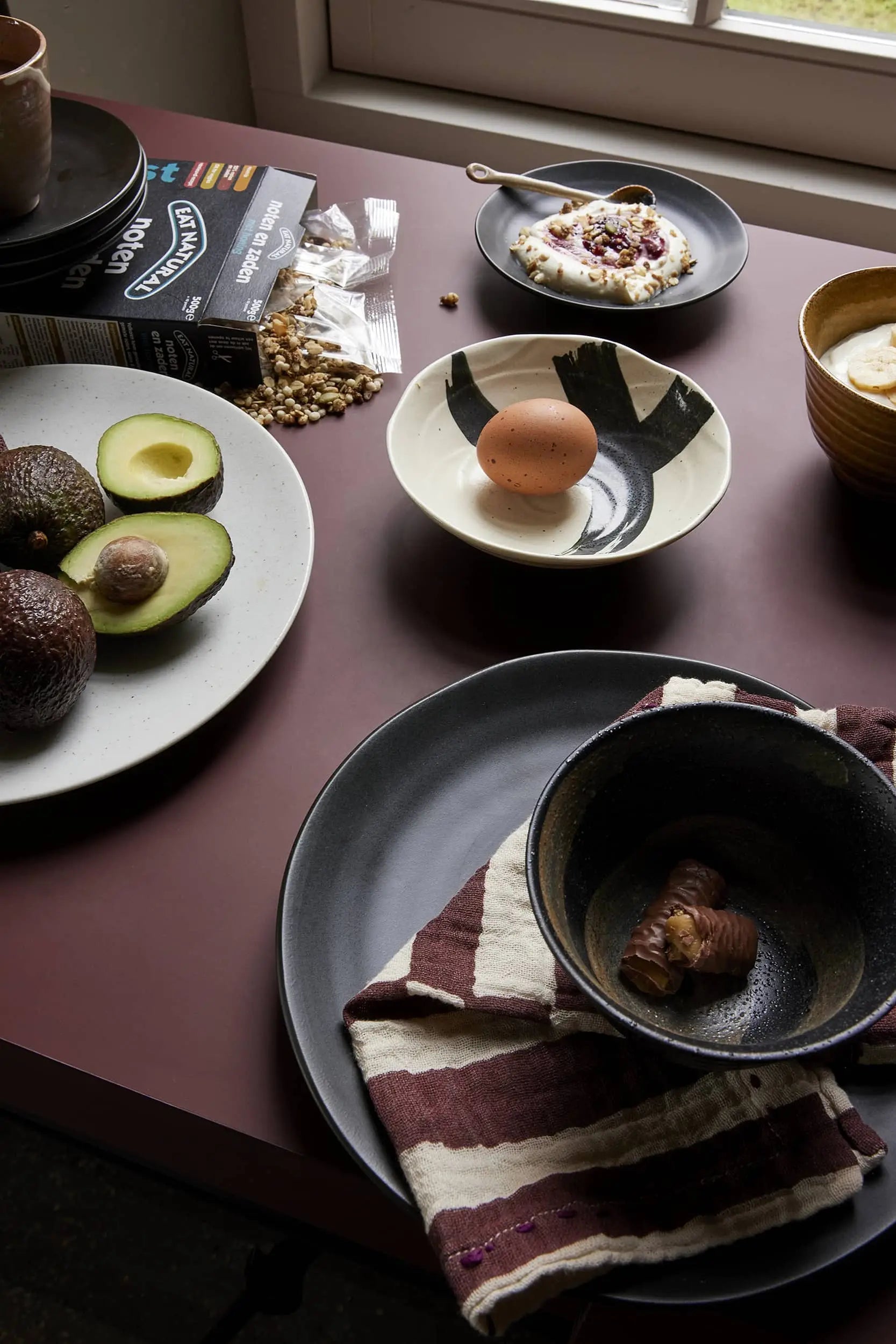 A mauve table set for breakfast features halved avocados, a brown egg in a black-and-white bowl, chestnuts, yogurt with toppings, granola, and HKLIVING Kyoto Ceramics Japanese Matcha Bowls (set of 3) beside a folded checked napkin on a plate.