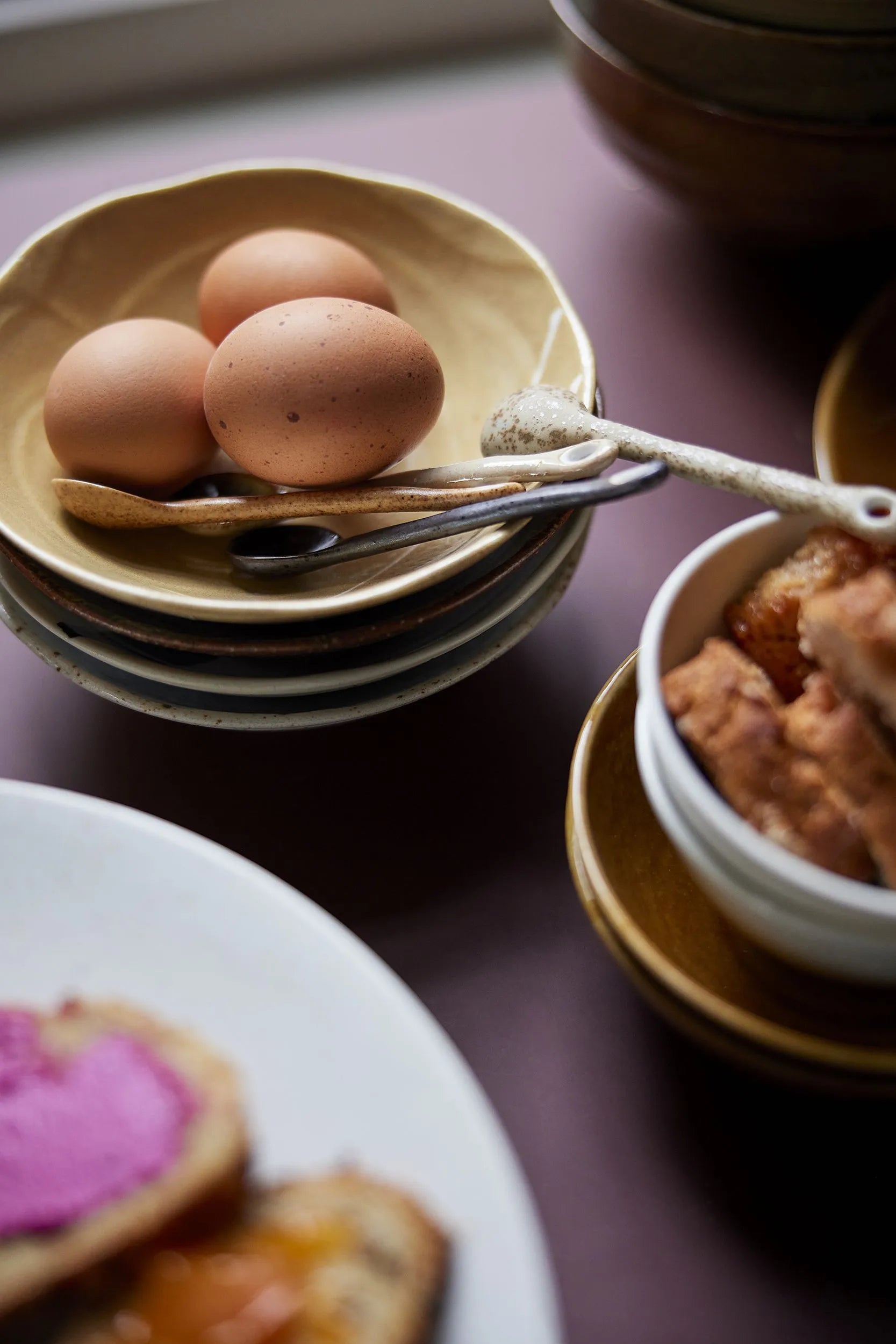 Three brown eggs in a yellow bowl sit beside an HKLIVING Kyoto Ceramics Japanese Tea Spoon, with stacked dishes and food nearby, capturing the charm of HKLIVING's Kyoto Ceramics.