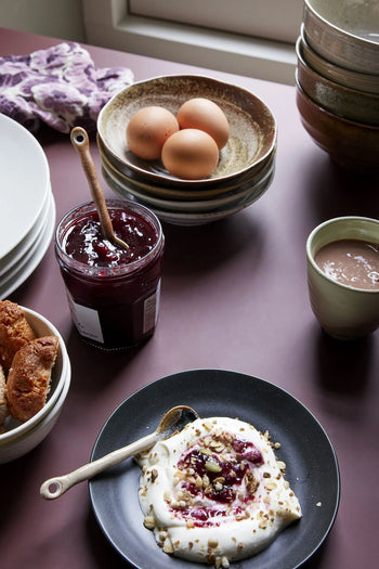 A breakfast scene with a black plate of yogurt, jam, nuts; croissants; HKLIVING Kyoto Ceramics bowls; brown eggs in a bowl; hot chocolate on mauve; and jam served with an HKLIVING Kyoto Ceramics Japanese Tea Spoon (Set of 4) by HKLIVING.