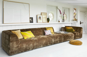 A long brown velvet sofa with colorful throw pillows and the HKliving Linen Cushion in mustard sits against a white wall decorated with art, photos, and decor. In front of the sofa are a small round table and matching floor cushion from HKliving.