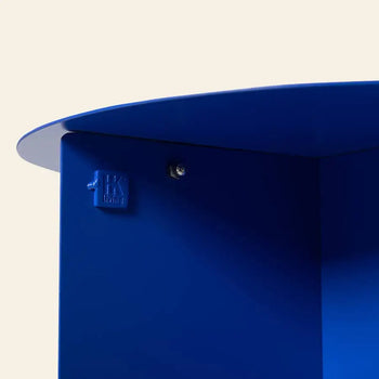 Close-up of the HKLIVING Metal Side Table Round in bright blue metal with smooth curves, a visible screw, and an embossed HKLIVING logo on the side, set against a light background.