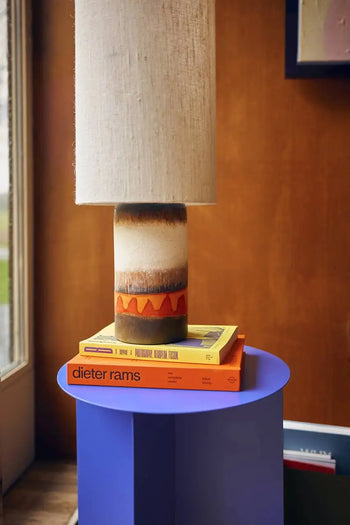 An HKLIVING Metal Side Table Round holds a ceramic table lamp with a beige shade on two stacked books—one orange, titled dieter rams, and one yellow—set against a wooden wall.