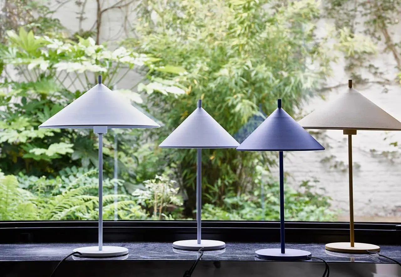 Four HKliving Metal Triangle Table Lamps M in pastel blue and beige by HKliving bring modern elegance to a windowsill, complementing the view of a lush garden. Designed for E14 Max 11w LED bulbs, they deliver both style and function.