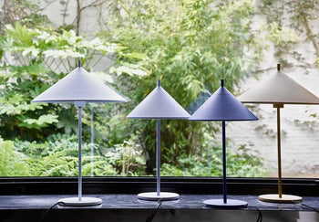 Four HKliving Metal Triangle Table Lamps M in pastel blue and beige by HKliving bring modern elegance to a windowsill, complementing the view of a lush garden. Designed for E14 Max 11w LED bulbs, they deliver both style and function.