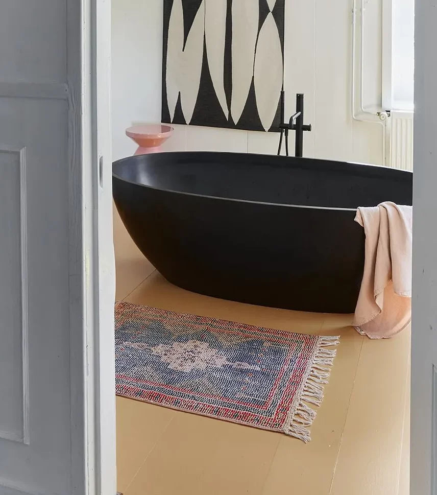 A modern bathroom features a black freestanding tub, a light pink towel, and the HKLIVING Printed Bathmat Overtufted by HKLIVING on the beige floor for comfort and style, with a pink bowl and abstract black-and-white wall art in the background.