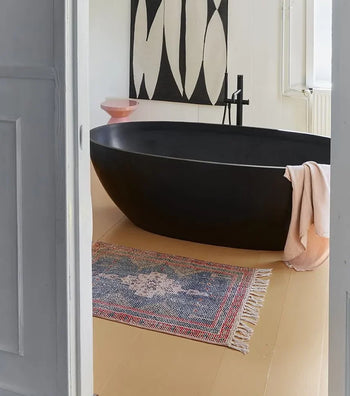 A modern bathroom features a black freestanding tub, a light pink towel, and the HKLIVING Printed Bathmat Overtufted by HKLIVING on the beige floor for comfort and style, with a pink bowl and abstract black-and-white wall art in the background.