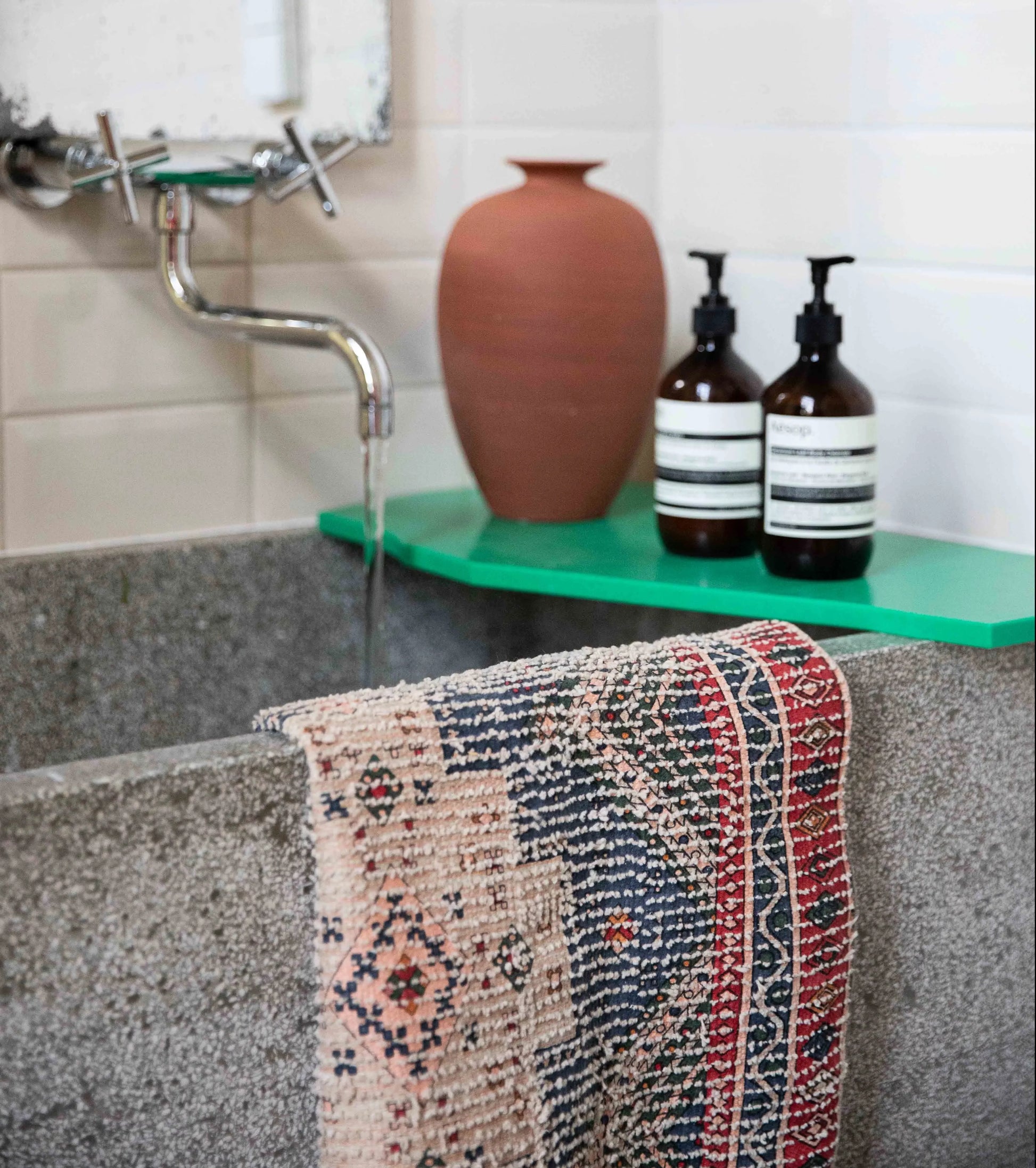 A gray stone sink with water running from a wall-mounted faucet, a patterned towel draped over the edge, and the HKLIVING Printed Bathmat Overtufted adds comfort underfoot; a green shelf behind holds a clay vase and two brown soap dispensers.
