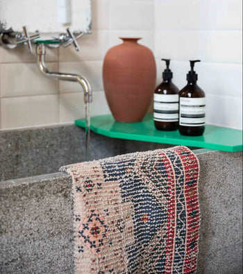 A gray stone sink with water running from a wall-mounted faucet, a patterned towel draped over the edge, and the HKLIVING Printed Bathmat Overtufted adds comfort underfoot; a green shelf behind holds a clay vase and two brown soap dispensers.