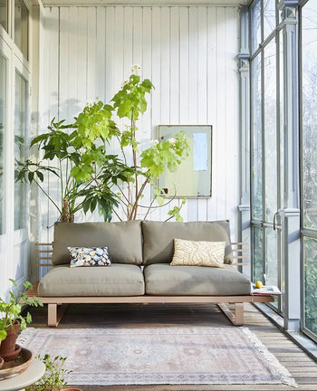 A cozy screened-in porch features a modern beige sofa, decorative pillows, leafy plant, and a small round side table with potted plants atop the eco-friendly HKLIVING Printed Indoor/Outdoor Rug (120x180) by HKLIVING on the wooden floor.