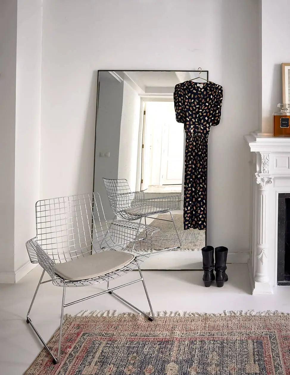 A minimalist room with a white wire chair, the HKLIVING Printed Rug Overtufted (150x240), a large floor mirror, and a black patterned dress with black boots placed near the white fireplace.