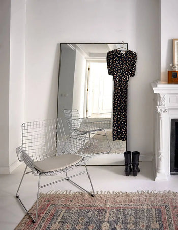 A minimalist room with a white wire chair, the HKLIVING Printed Rug Overtufted (150x240), a large floor mirror, and a black patterned dress with black boots placed near the white fireplace.