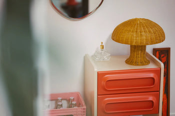 A small corner features a retro dresser with red drawers, the HKLIVING Rattan Table Lamp in yellow mushroom shape adding cozy light, a glass perfume bottle, and a pink crate with bottles, all set against a white wall partly reflected in a mirror.