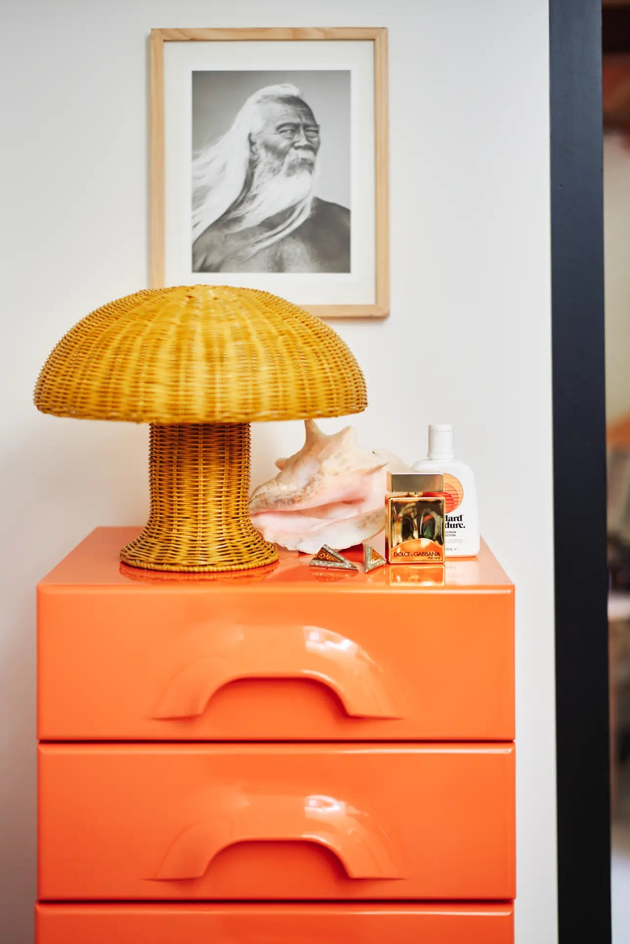 A vibrant orange cabinet holds the HKLIVING Rattan Table Lamp, perfume bottles, a large seashell, and lotion. Above it hangs a framed black-and-white photo of a long-haired bearded man.
