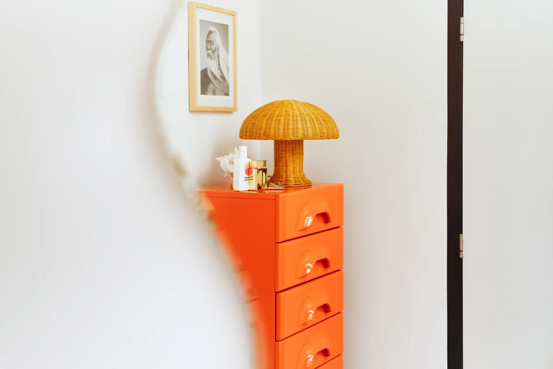 A modern room features a bold orange drawer unit and cozy lighting from the HKLIVING Rattan Table Lamp. Small decor accents the space, while a black-and-white portrait hangs above, partially blurred by a curved object in front.