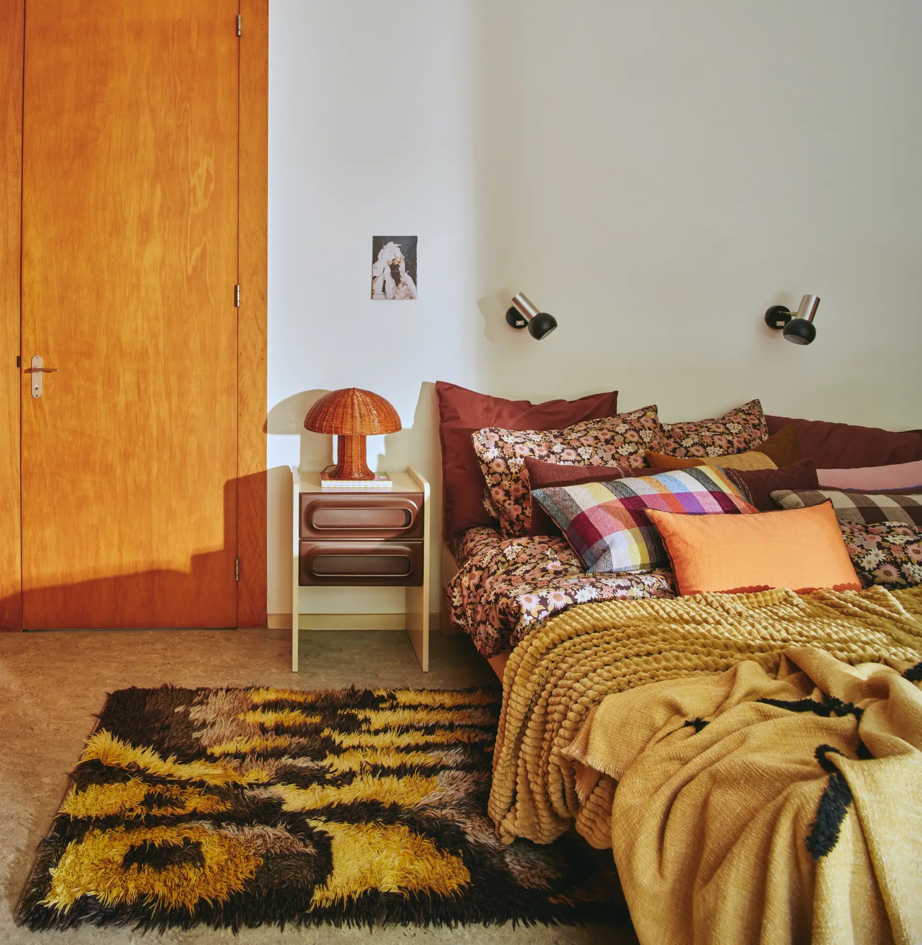 A cozy bedroom features a bed with patterned pillows and blankets, a small nightstand topped with the HKLIVING Rattan Table Lamp by HKLIVING, wall-mounted lights, a fuzzy rug, and a closed wooden door. Warm, earthy tones and cozy lighting set the mood.