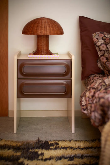 A two-drawer brown bedside table displays the HKLIVING Rattan Table Lamp, casting warm, cozy light over a book. The bed beside it features a floral blanket and brown pillow, all set atop a patterned rug.