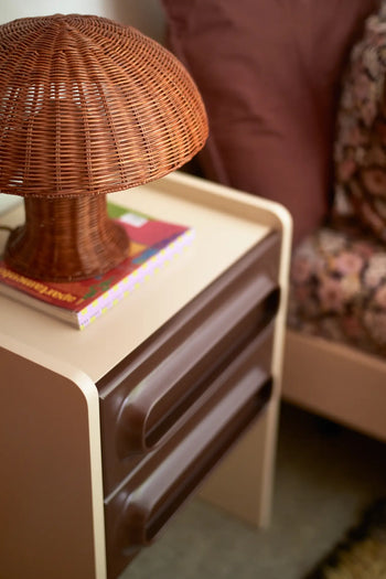 A close-up of a nightstand with two brown drawers, the HKLIVING Rattan Table Lamp by HKLIVING shaped like a mushroom giving cozy lighting, and a stack of books. Beside it is a bed with pink and floral bedding.