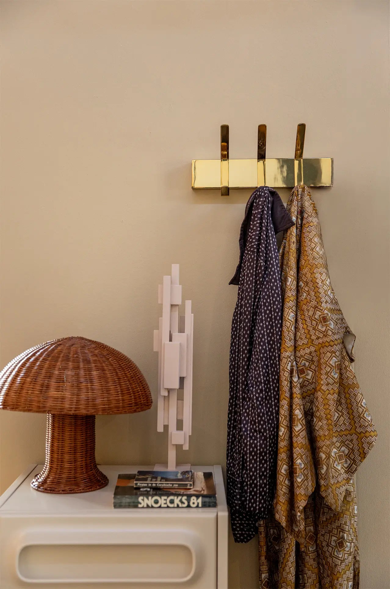 The HKLIVING Rattan Table Lamp and stacked books rest on a white table beside a brass wall hook with two patterned scarves, casting cozy light against a beige wall.