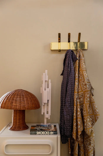 The HKLIVING Rattan Table Lamp and stacked books rest on a white table beside a brass wall hook with two patterned scarves, casting cozy light against a beige wall.
