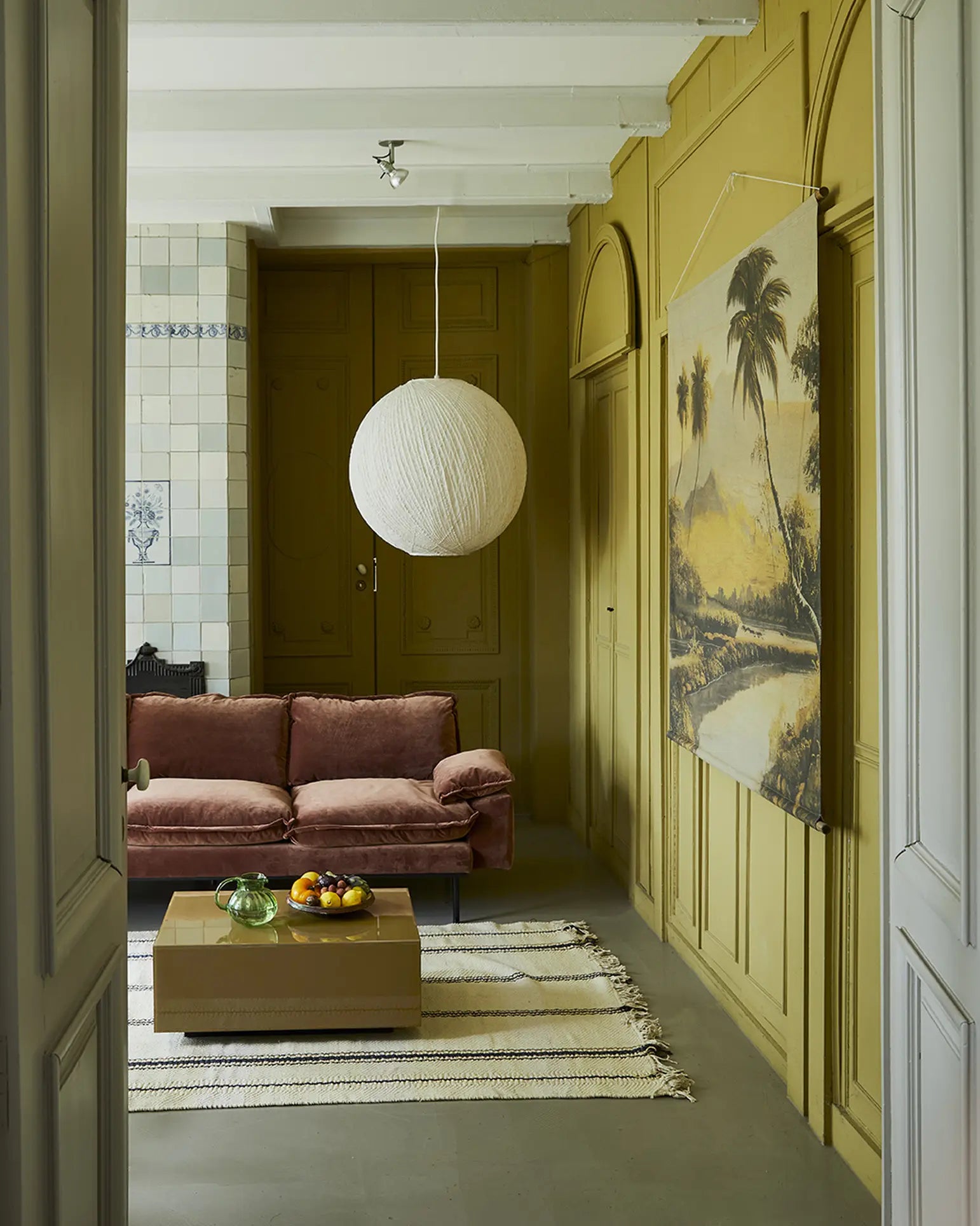 A cozy living room with mustard yellow paneled walls, a mauve HKLIVING Retro Sofa (2-seater), tan coffee table with fruit bowls, striped rug, large paper lantern light, tiled wall accents, and a tropical landscape painting.