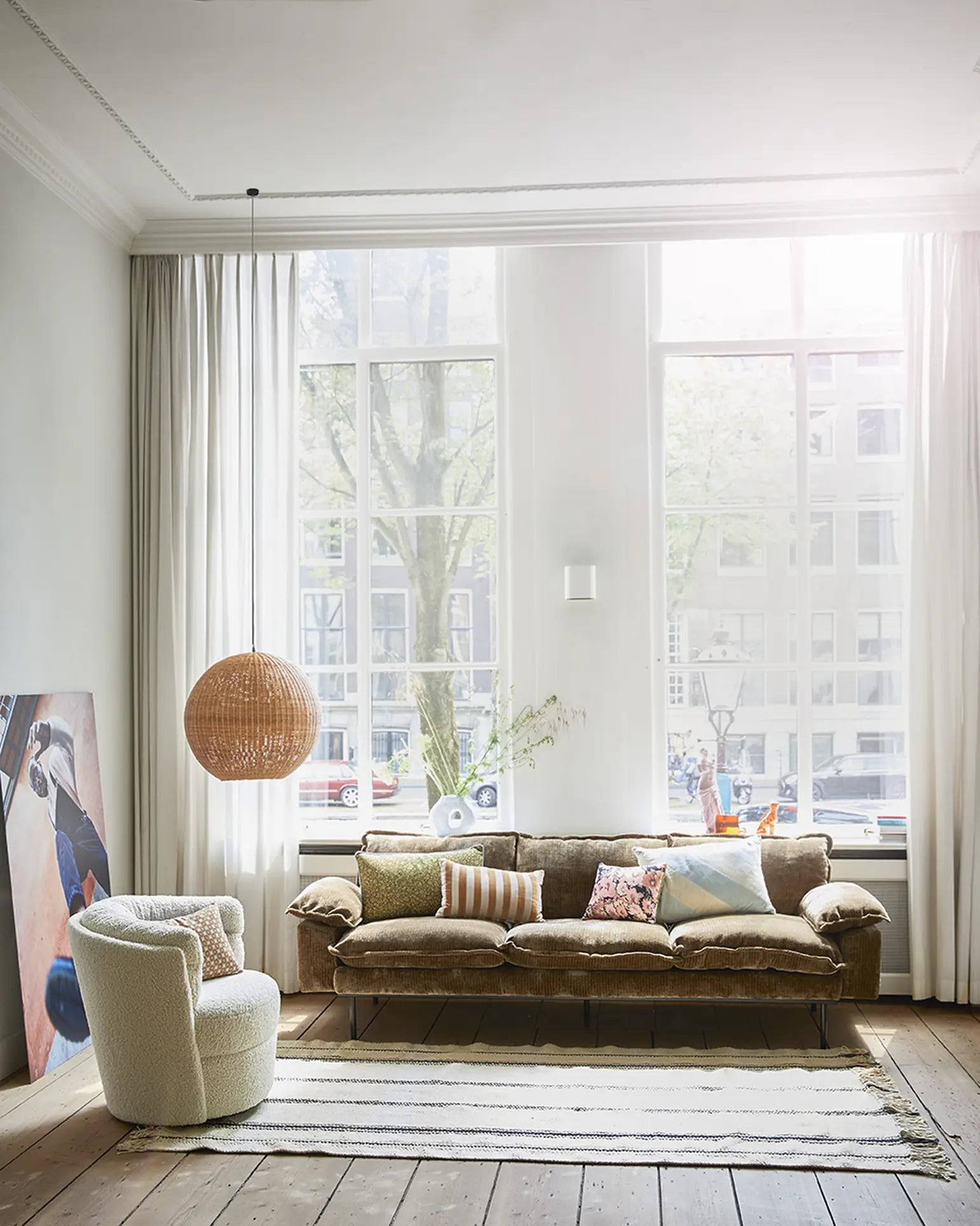 Bright living room with tall windows, sheer white curtains, HKLIVING Retro Sofa (3-Seats) by HKLIVING with assorted pillows, round white chair, striped rug, large wicker hanging lamp, and abstract painting leaning against the wall.