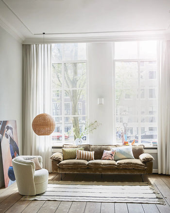 Bright living room with tall windows, sheer white curtains, HKLIVING Retro Sofa (3-Seats) by HKLIVING with assorted pillows, round white chair, striped rug, large wicker hanging lamp, and abstract painting leaning against the wall.