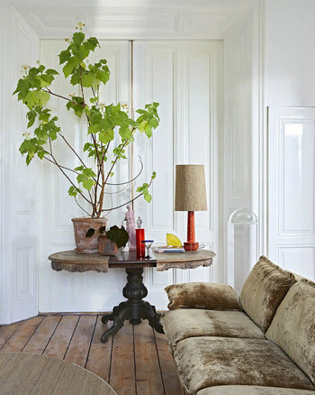 A cozy living room featuring the HKLIVING Retro Sofa, 3-Seats by HKLIVING, a round wooden table with a tall potted plant, a red lamp, and decor, set against white paneled walls and wood floorboards.