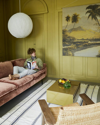 A person relaxes on the HKLIVING Retro Sofa, 4-Seats, in soft pink velvet, reading a magazine in a mustard yellow room with paneled walls, a globe pendant light, tropical art, striped rug, gold coffee table, and wicker chair.