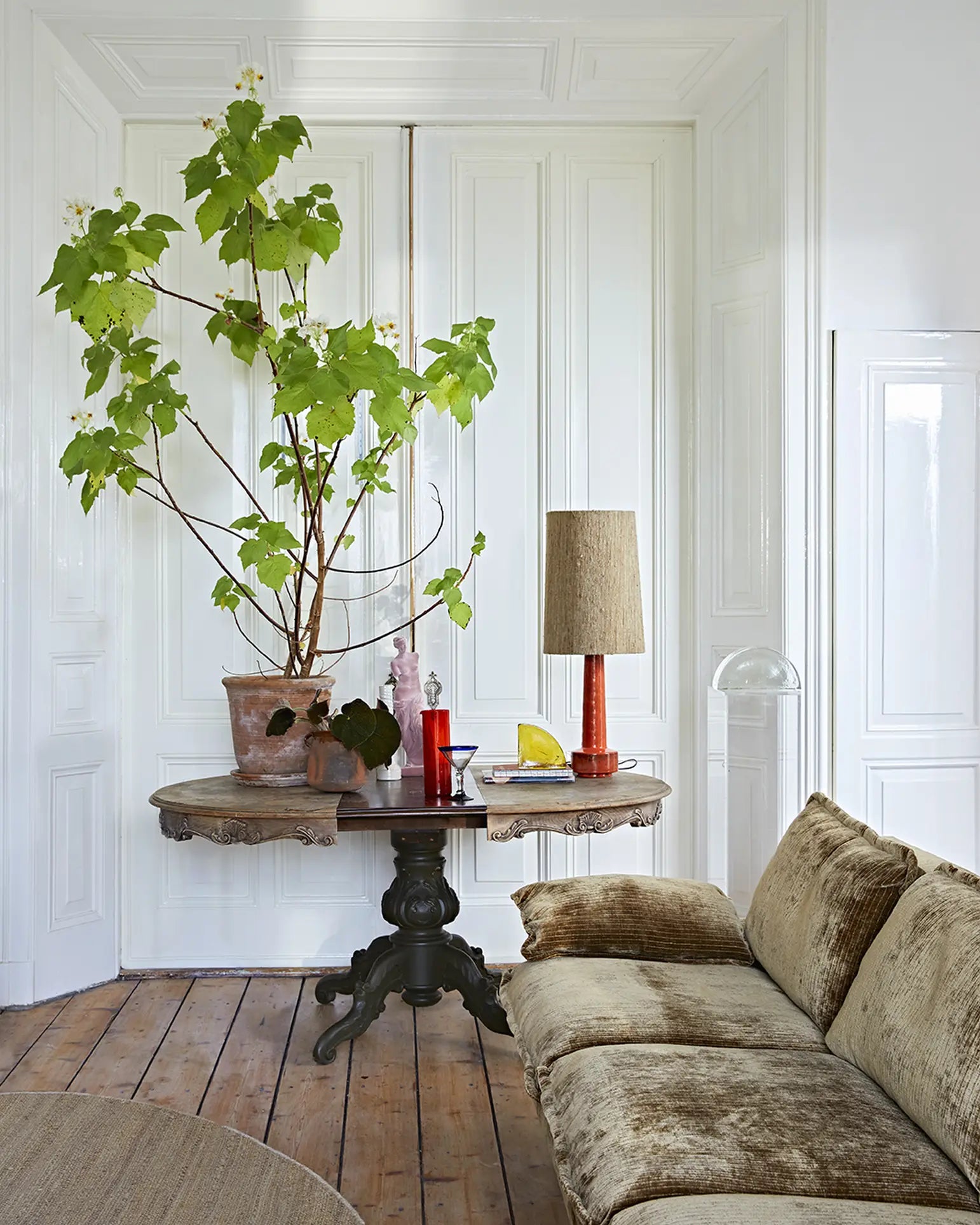 A cozy living room featuring the HKLIVING Retro Sofa, 4-Seats, a round wooden table with a potted plant, two vases, a red glass, a yellow object, and a lamp—all set against white paneled walls and a wooden floor.