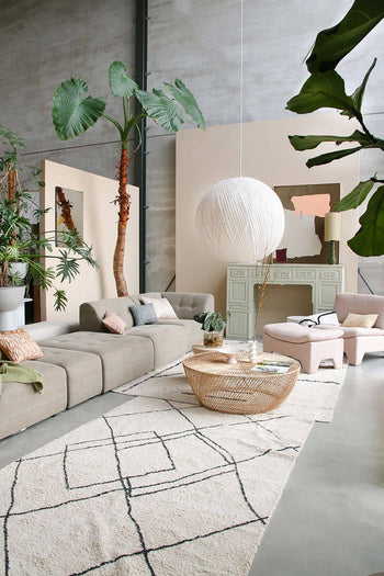 A modern living room with neutral furniture, leafy plants, a round wicker coffee table, geometric rug, cosy HKliving Soft Woven Throw With Black Tufted Lines for texture, pale green sideboard, white pendant light, high ceilings, and soft natural light.
