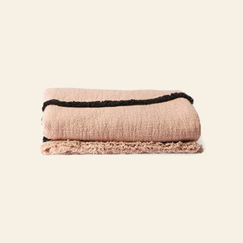 The HKliving Soft Woven Throw With Black Tufted Lines is a cosy cotton blanket in pink, featuring black stripes near the edge and a stylish texture, folded neatly on a plain, light-colored background.