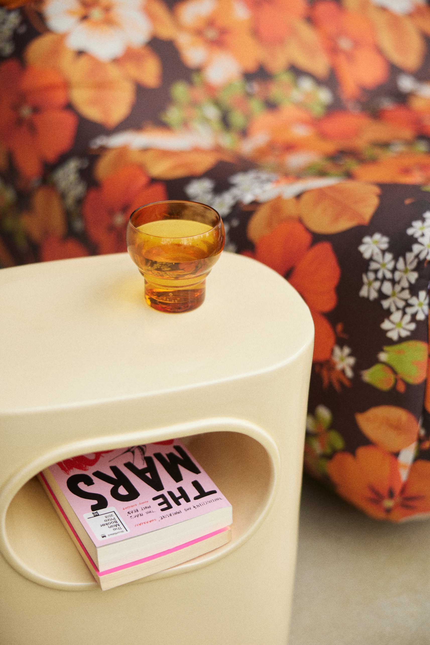 The HKLIVING Space Side Table by HKLIVING, in beige, offers built-in storage and is shown with an orange glass cup and a pink book titled THE MARS. Behind it sits a vibrant sofa with an orange and floral pattern.