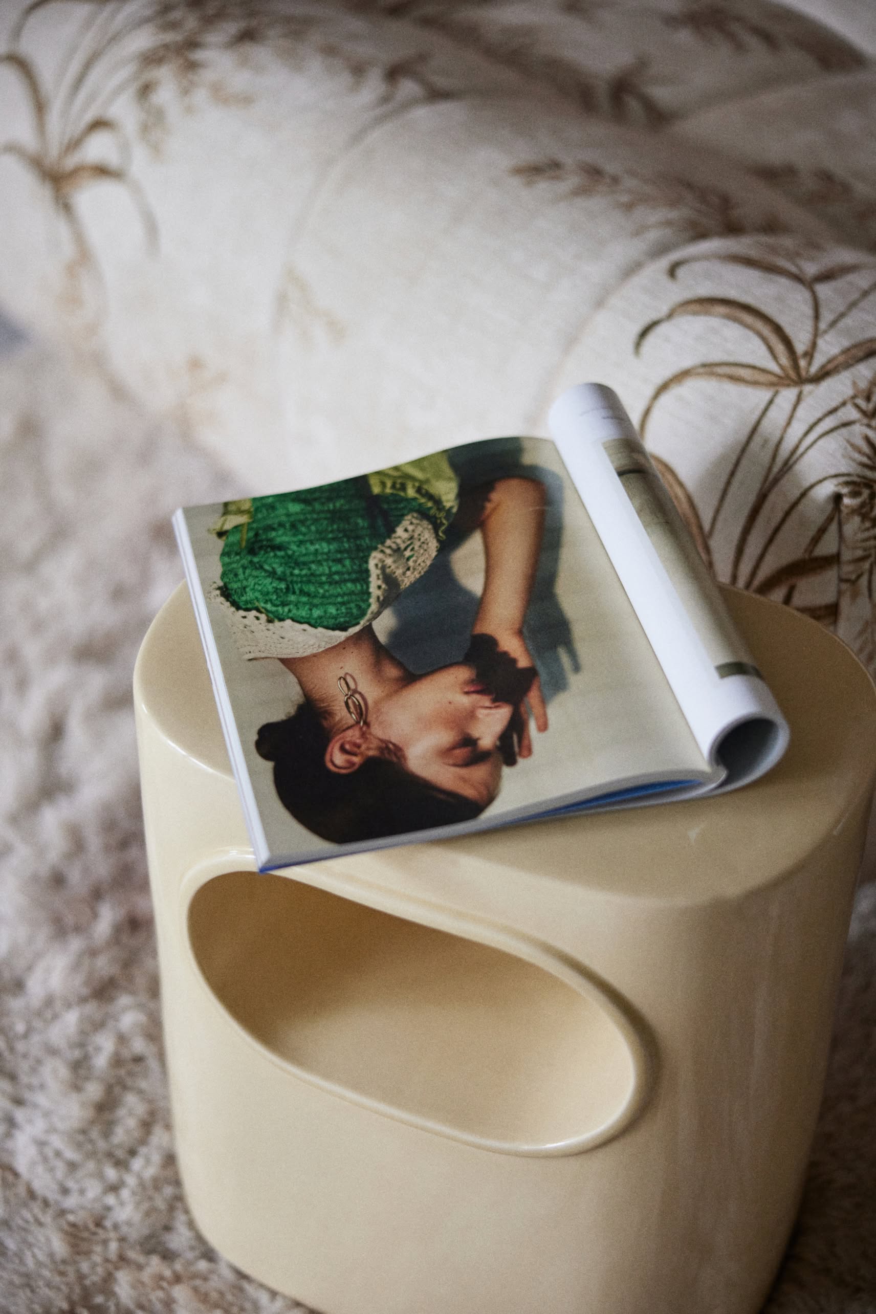 An open magazine rests on the HKLIVING Space Side Table with built-in storage by HKLIVING, set beside a patterned cream couch and textured carpet—an ideal addition to contemporary interiors.
