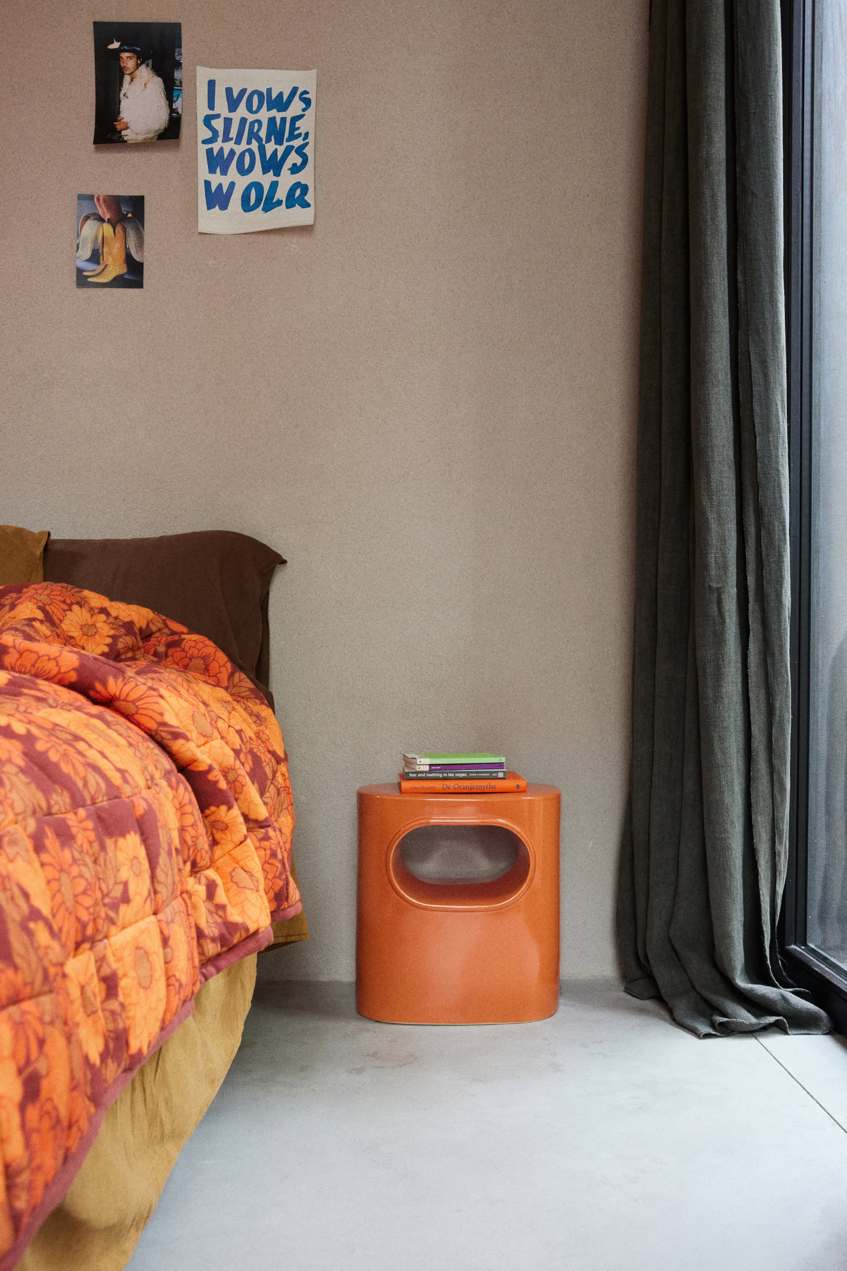 A cozy bedroom corner with an orange floral-patterned bedspread, an HKLIVING Space Side Table topped with books, green curtains, and three art prints or photos displayed on the beige wall above.