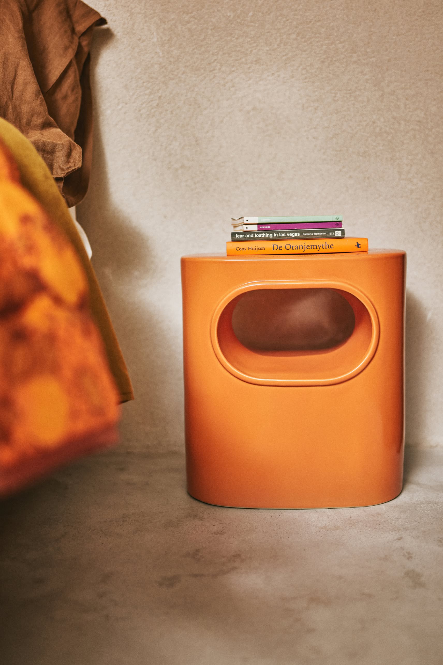 The HKLIVING Space Side Table, an orange oval piece with a cut-out handle and built-in storage, holds books beside a bed with orange and brown bedding. Its modern design by HKLIVING fits perfectly on a light floor in any contemporary space.