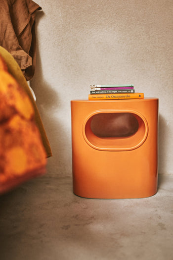 The HKLIVING Space Side Table, an orange oval piece with a cut-out handle and built-in storage, holds books beside a bed with orange and brown bedding. Its modern design by HKLIVING fits perfectly on a light floor in any contemporary space.