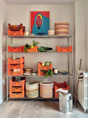 A metal shelving unit with orange crates, baskets, dishes, kitchenware, and fresh produce stands on a wooden floor. Above hangs colorful portrait art. The HKLIVING Stoneware Side Table - Chrome by HKLIVING sits near a silver trash bin.