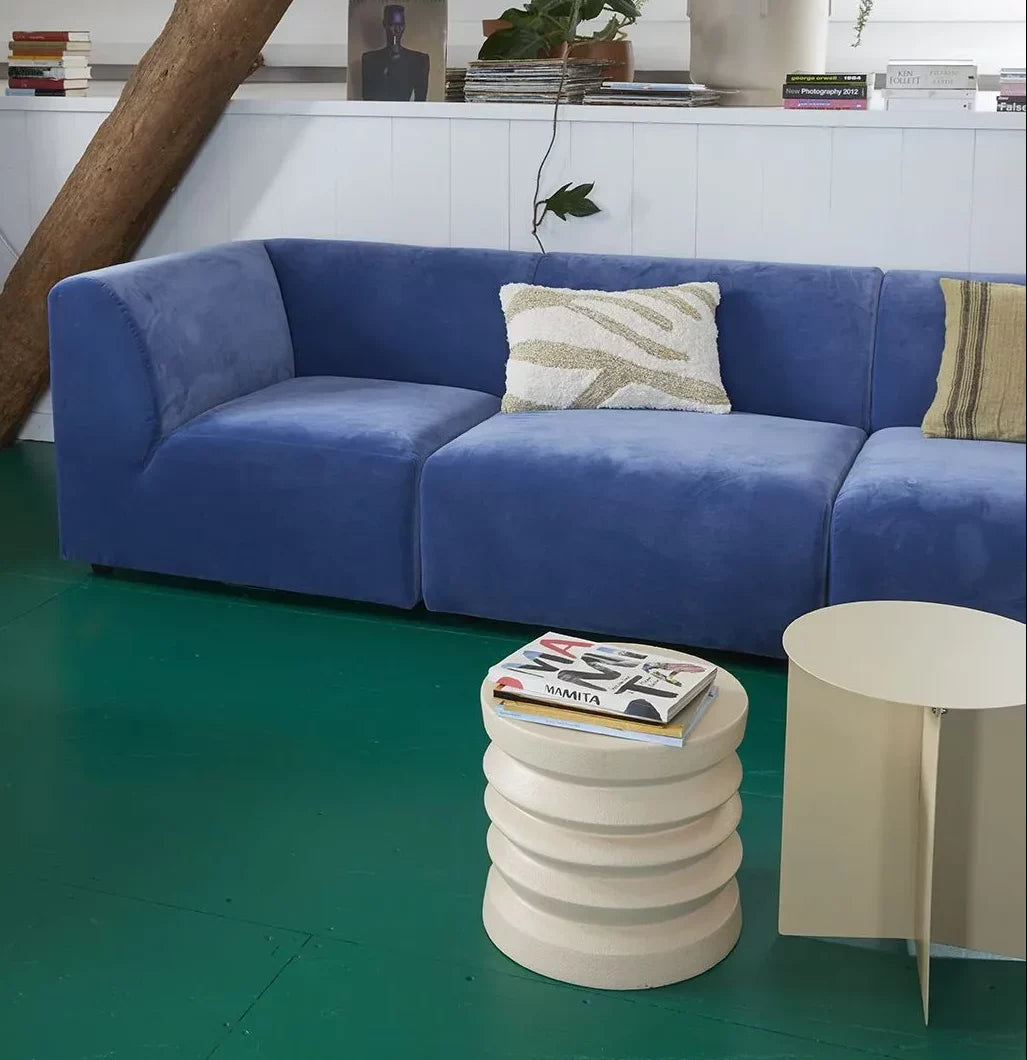 A modern living room features a blue velvet sofa, a decorative pillow, the HKLIVING Stoneware Side Table by HKLIVING with a magazine on top, a round beige table, bright green flooring, and books and plants on the shelf behind the sofa.