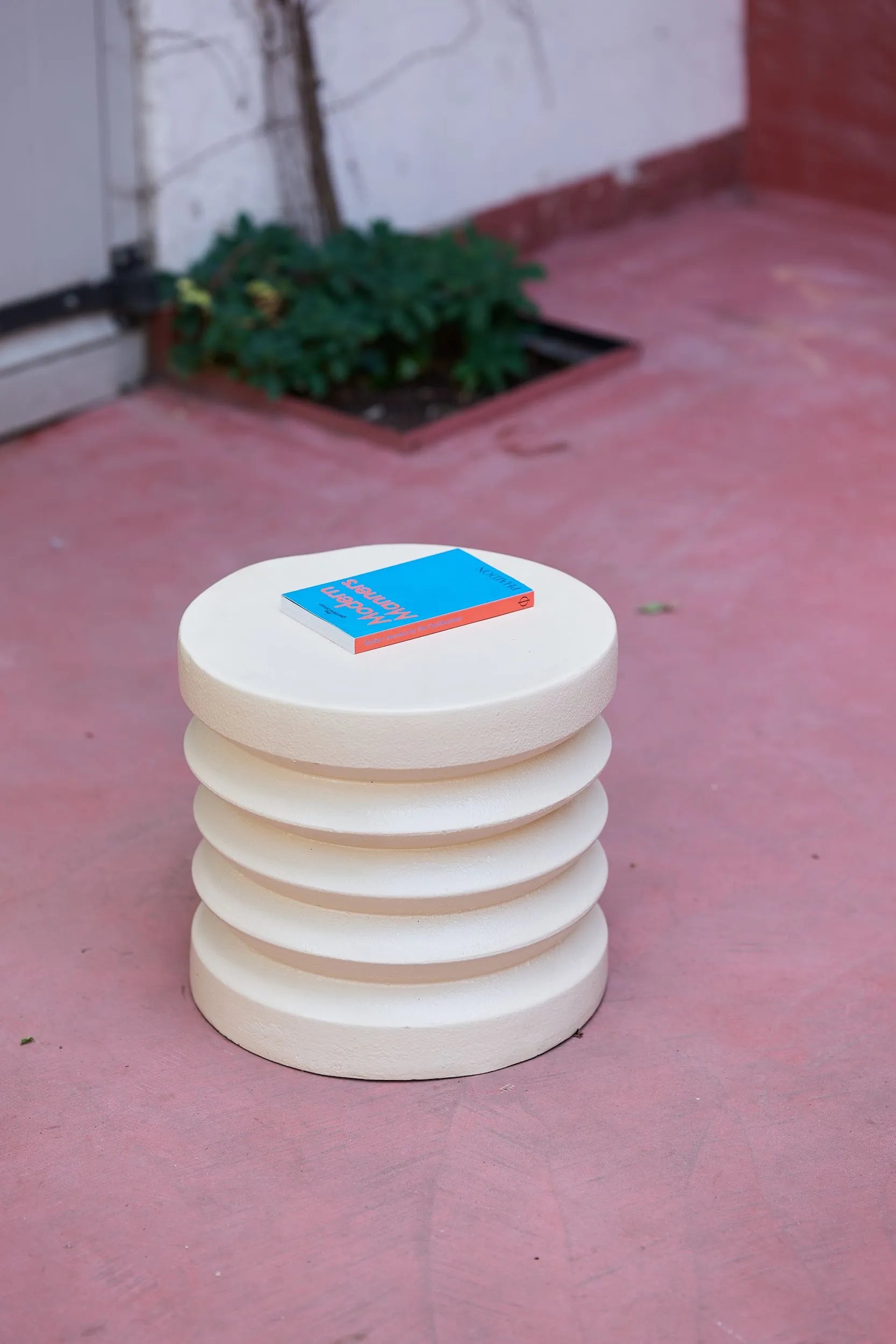 The HKLIVING Stoneware Side Table by HKLIVING, round and ribbed in white, stands on a pinkish patio floor. A blue and orange book lies atop its durable surface, with a small green plant nearby in a ground-level planter.