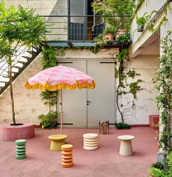 A colorful patio with pink flooring features a pink-and-yellow umbrella, pastel stools, plants, a pink bench, and the HKLIVING Stoneware Side Table by HKLIVING. A staircase leads to an upper level with extra greenery and potted plants.