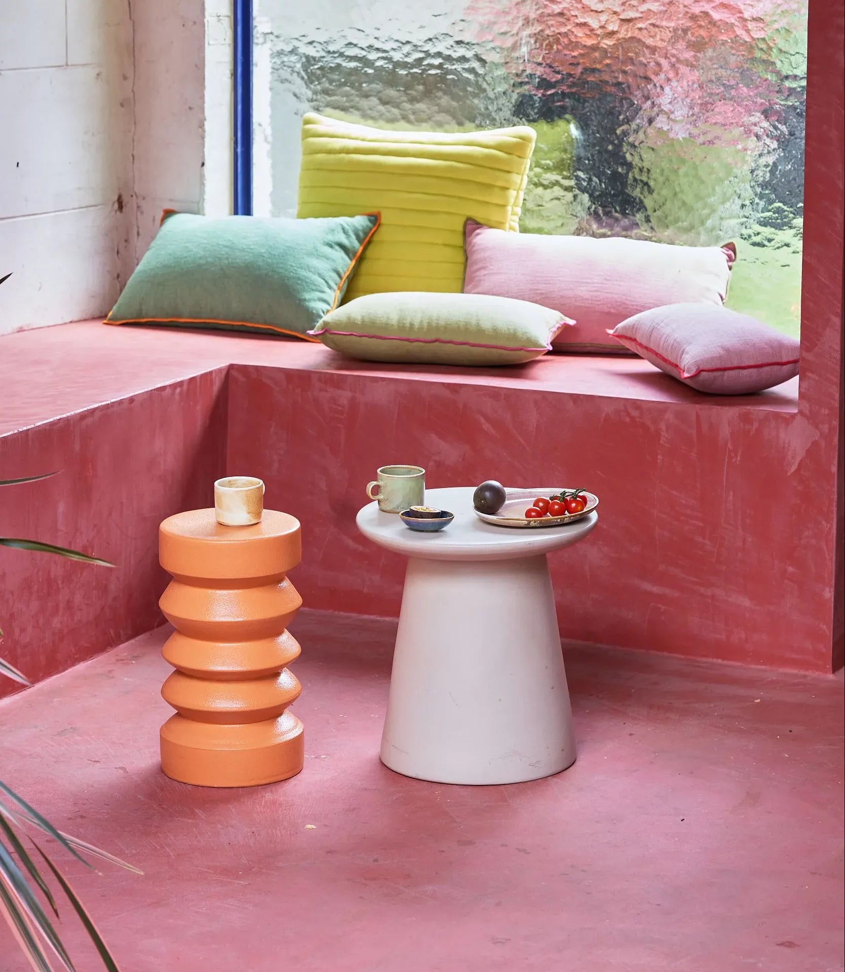 A pink seating nook with pastel cushions, an HKLIVING Stoneware Side Table by HKLIVING holding a cup and plate of food, and an orange sculptural stool; a large textured window lets in light.